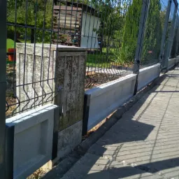 Długi fragment ogrodzenia panelowego z podmurówką betonową i słupkami, widok z boku, na pierwszym planie skrzynka elektryczna na słupku.