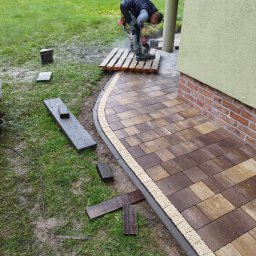Usługi Remontowo-Budowlane Łukasz Bąk - Pracownik tnie kostkę brukową na palecie, podczas układania ścieżki z kostki brukowej w ogrodzie. Częściowo ułożona ścieżka z obrzeżami. W tle zielony trawnik.