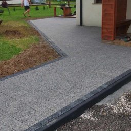 Usługi Remontowo-Budowlane Łukasz Bąk - Ścieżka z szarej kostki brukowej z czarnym obramowaniem, prowadząca do domu w Łodygowicach. Widoczny fragment ogrodu i elewacji budynku.