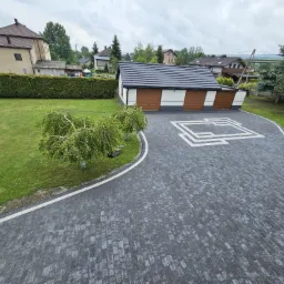 Szara kostka brukowa ułożona na podjeździe z białymi akcentami, widok z góry, w tle budynek garażu z brązowymi bramami i zadbany ogród z zielonym trawnikiem.