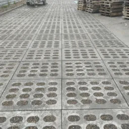 Szara ażurowa nawierzchnia z wypełnieniem żwirowym, układana w regularny wzór, z widoczną przyczepą i stertą palet w tle, sugerująca parking lub drogę dojazdową.