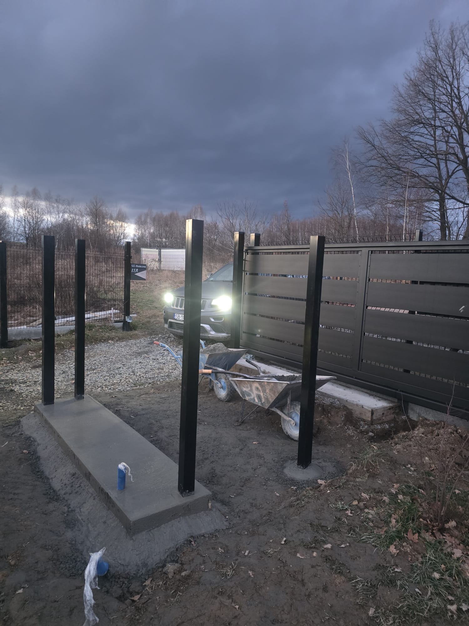 Montaż słupków ogrodzeniowych z betonową podstawą i przygotowaną bramą przesuwną na posesji, widoczny wózek budowlany i samochód z włączonymi światłami w tle, pochmurne niebo.