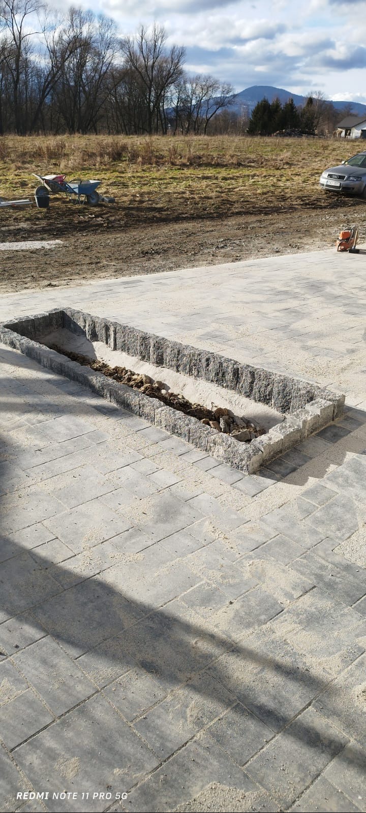 Kwadratowa rabata z kostki granitowej w trakcie budowy, otoczona ułożonym brukiem, w tle krajobraz z drzewami i wzgórzami.