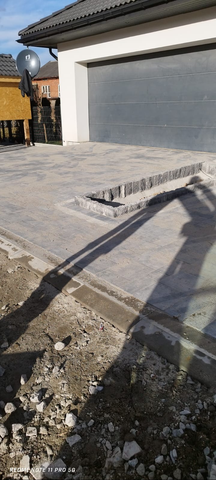 Ułożona szara kostka brukowa na podjeździe z obrzeżami, widoczny fragment domu z szarą bramą garażową i satelitą, teren wokół w trakcie prac ziemnych.