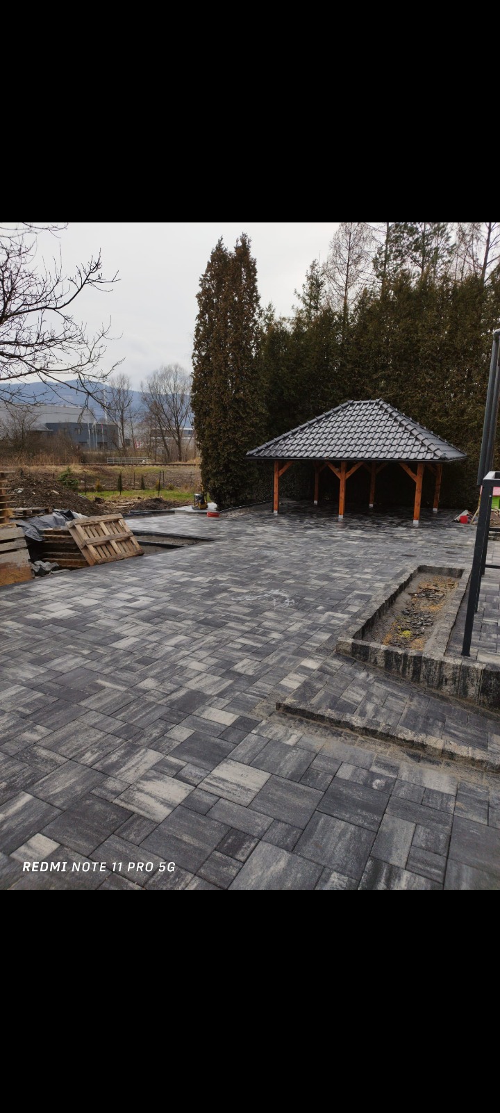 Ułożona szara kostka brukowa na posesji z drewnianą altaną w tle, widoczne palety z materiałami budowlanymi i fragment ogrodu.