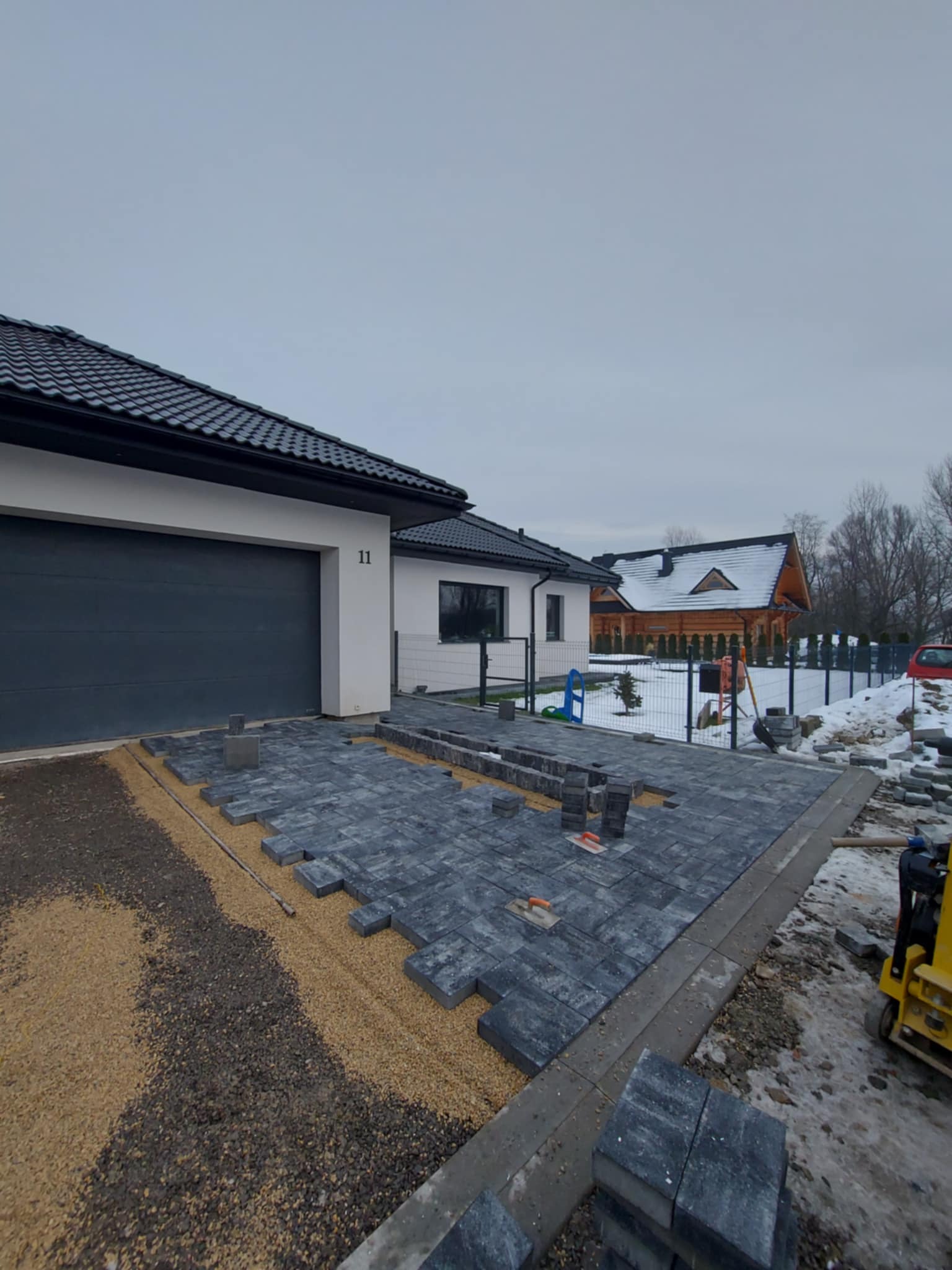 Częściowo ułożona, szara kostka brukowa przed nowoczesnym domem z ciemną bramą garażową i numerem 11, widoczny ubijak i podsypka z piasku.