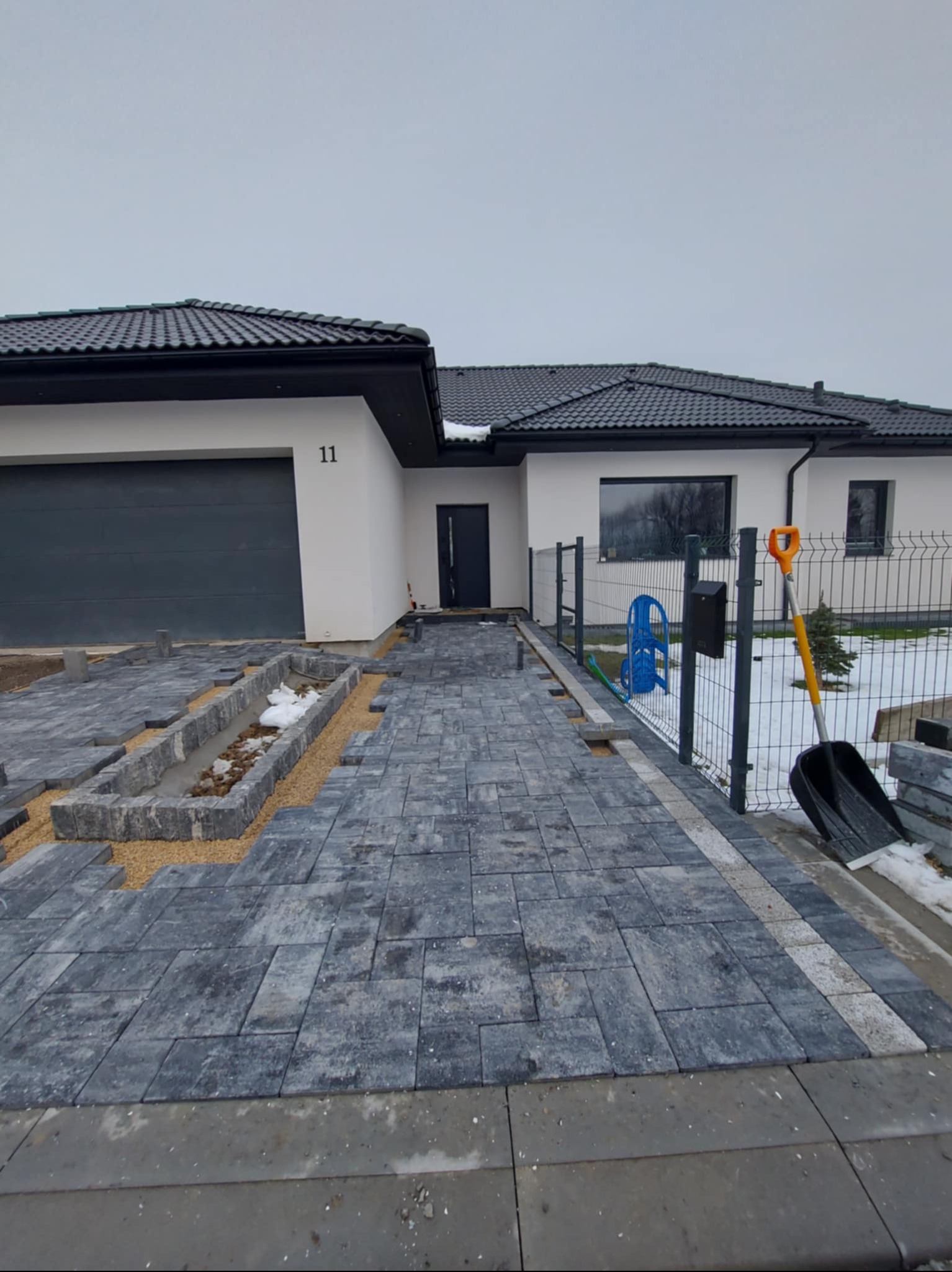 Niedokończona ścieżka z kostki brukowej prowadząca do nowoczesnego domu z numerem 11. Widoczny fragment ogrodu, narzędzia i częściowo ułożona kostka.