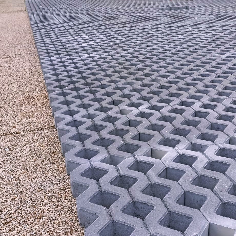 Szara ażurowa kostka brukowa o zygzakowatym wzorze, ułożona na powierzchni obok żwirowego podłoża. Widok z góry ukazuje regularny układ elementów.