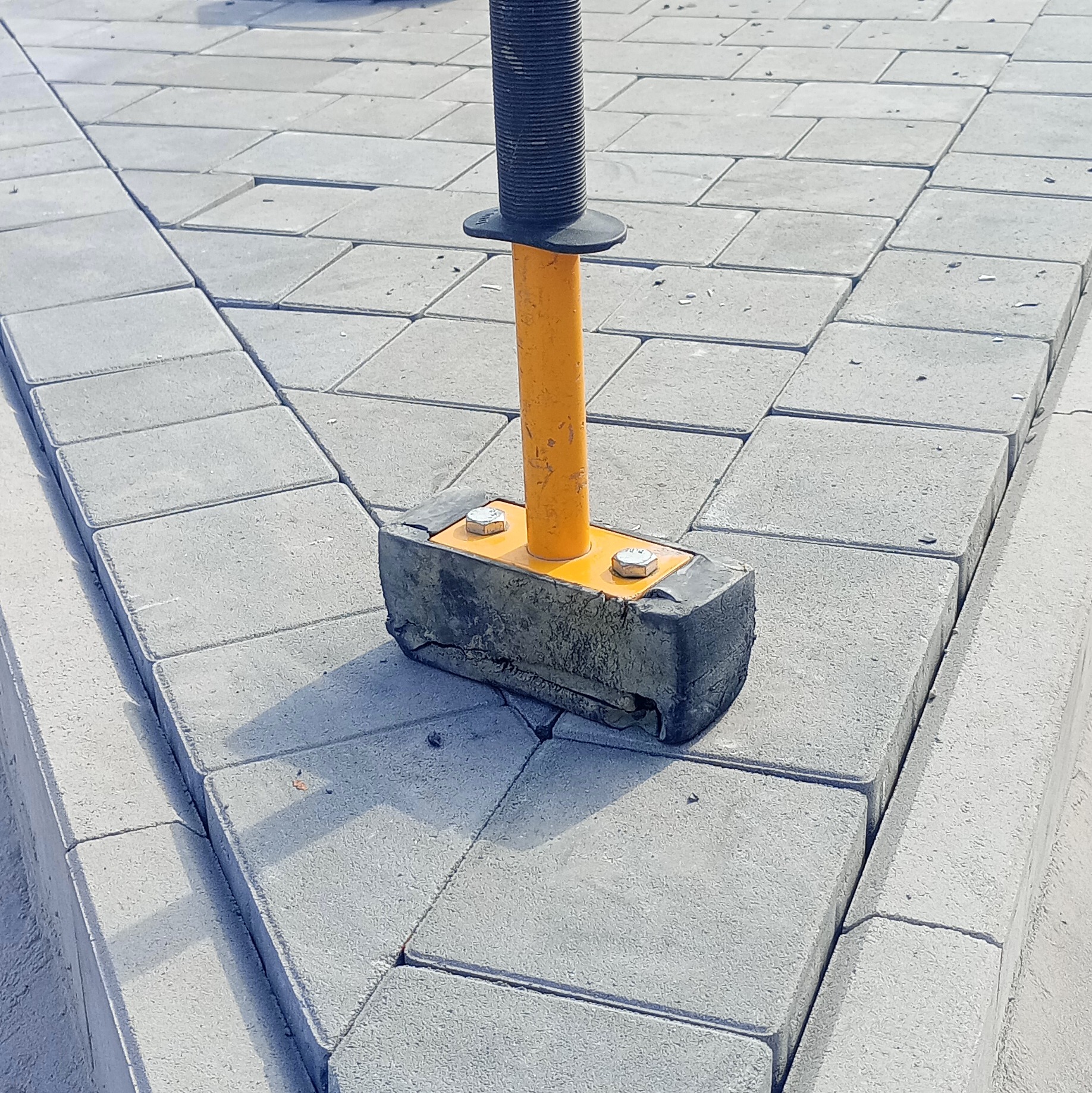 Szare kostki brukowe ułożone pod kątem prostym, z gumowym młotkiem z żółtą rączką na środku. Widoczne krawężniki i regularny wzór ułożenia.
