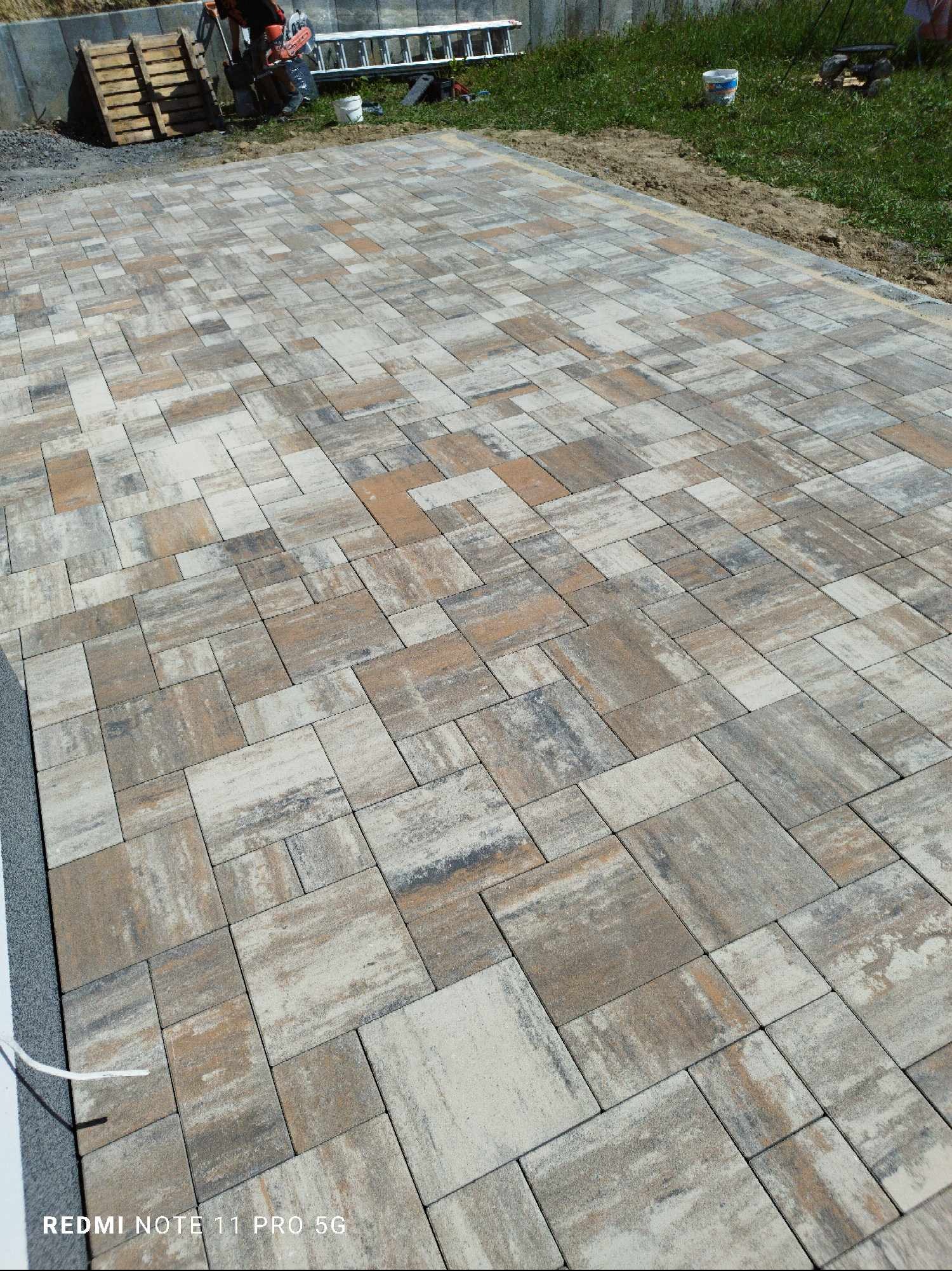 Ułożona kostka brukowa w odcieniach beżu i szarości, widok z góry, z paletą i narzędziami w tle. Nietypowy wzór ułożenia kostki.