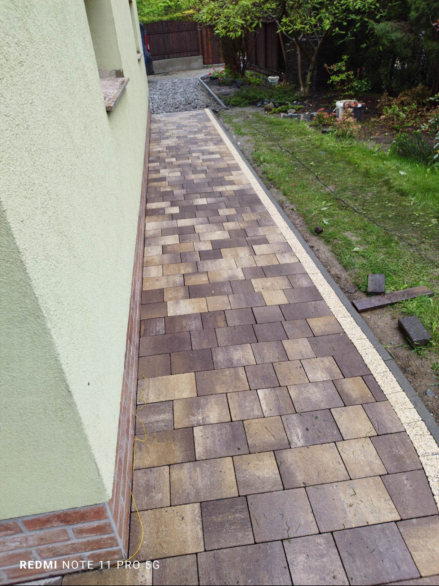 Ścieżka z kostki brukowej w odcieniach brązu i beżu, obrzeża z jasnej kostki, trawnik z boku, ściana budynku z oknem w tle, widok z góry.