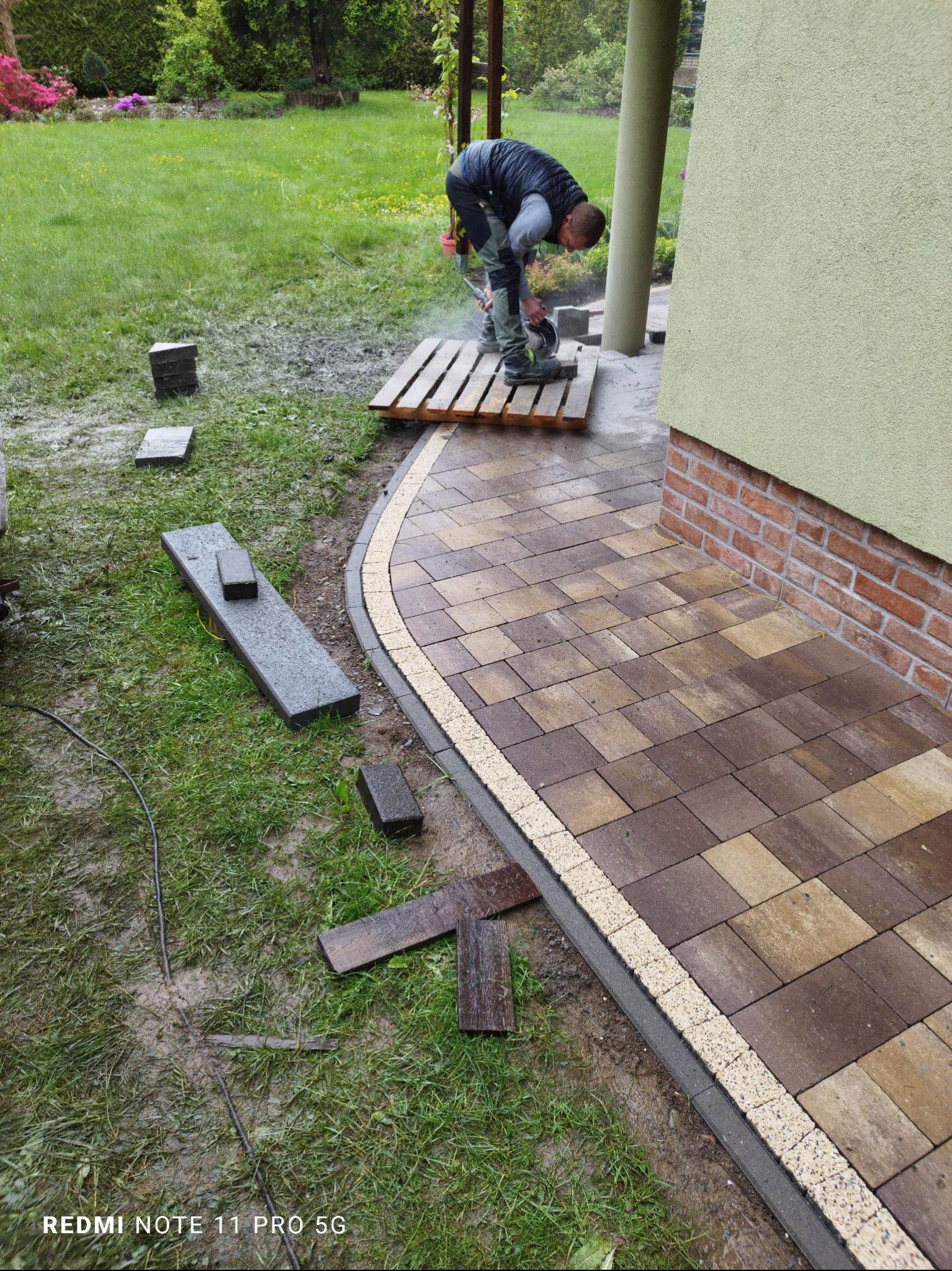 Pracownik tnie kostkę brukową na palecie, podczas układania ścieżki z kostki brukowej w ogrodzie. Częściowo ułożona ścieżka z obrzeżami. W tle zielony trawnik.