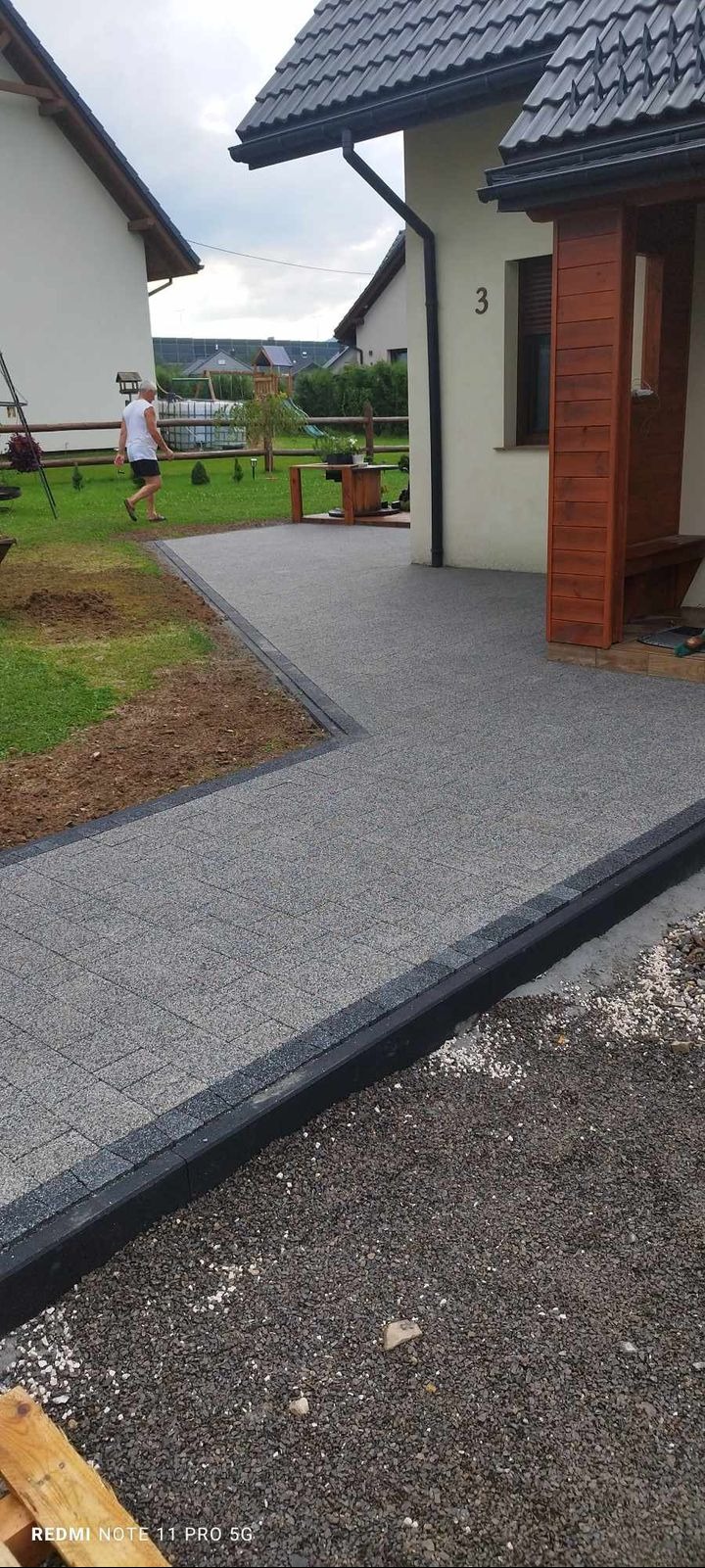 Ścieżka z szarej kostki brukowej z czarnym obramowaniem, prowadząca do domu w Łodygowicach. Widoczny fragment ogrodu i elewacji budynku.