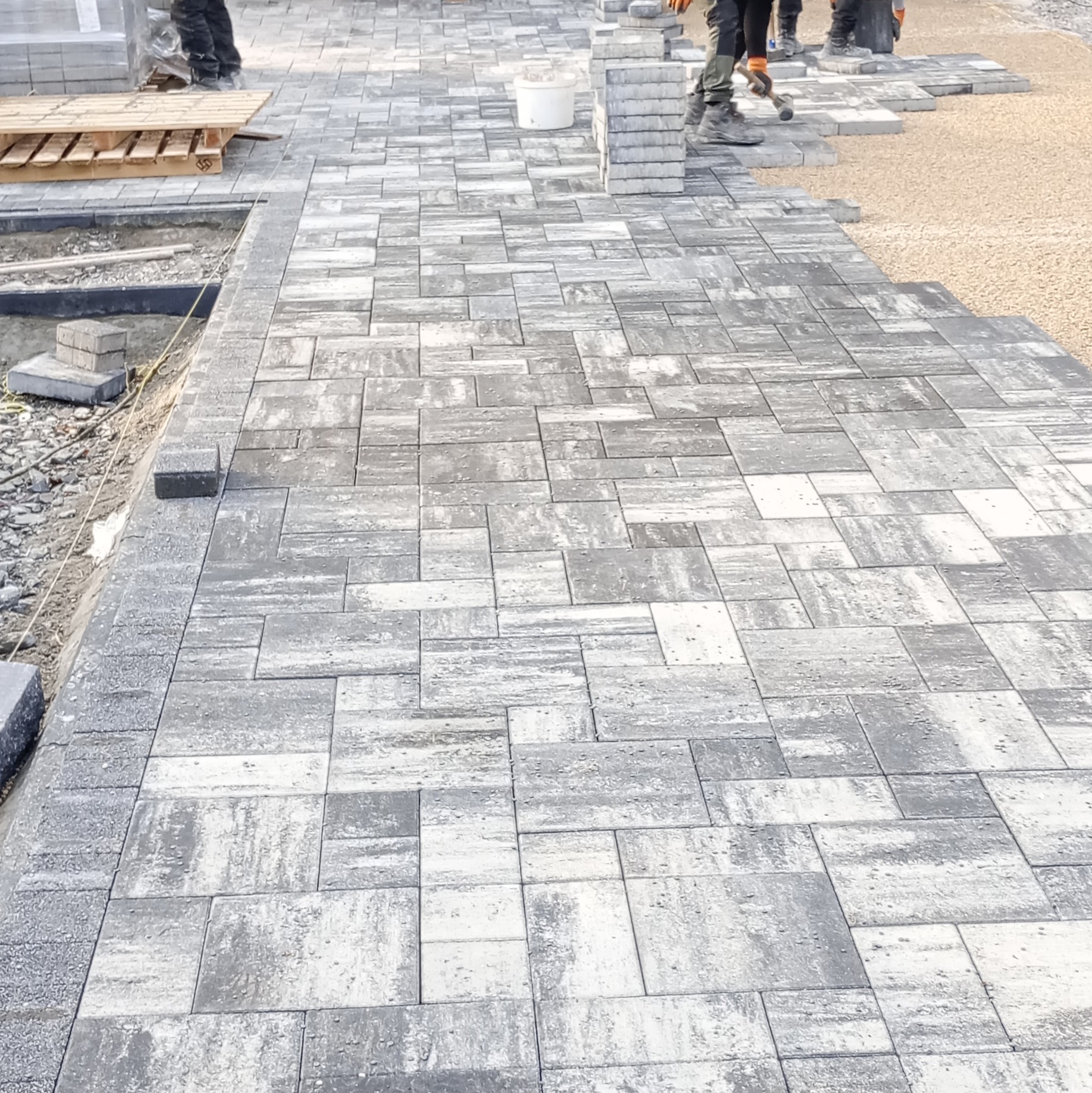 Układanie szarej kostki brukowej o nieregularnym wzorze. W tle paleta z kostką i pracownicy w trakcie pracy. Widoczny fragment podłoża przygotowanego pod kostkę.
