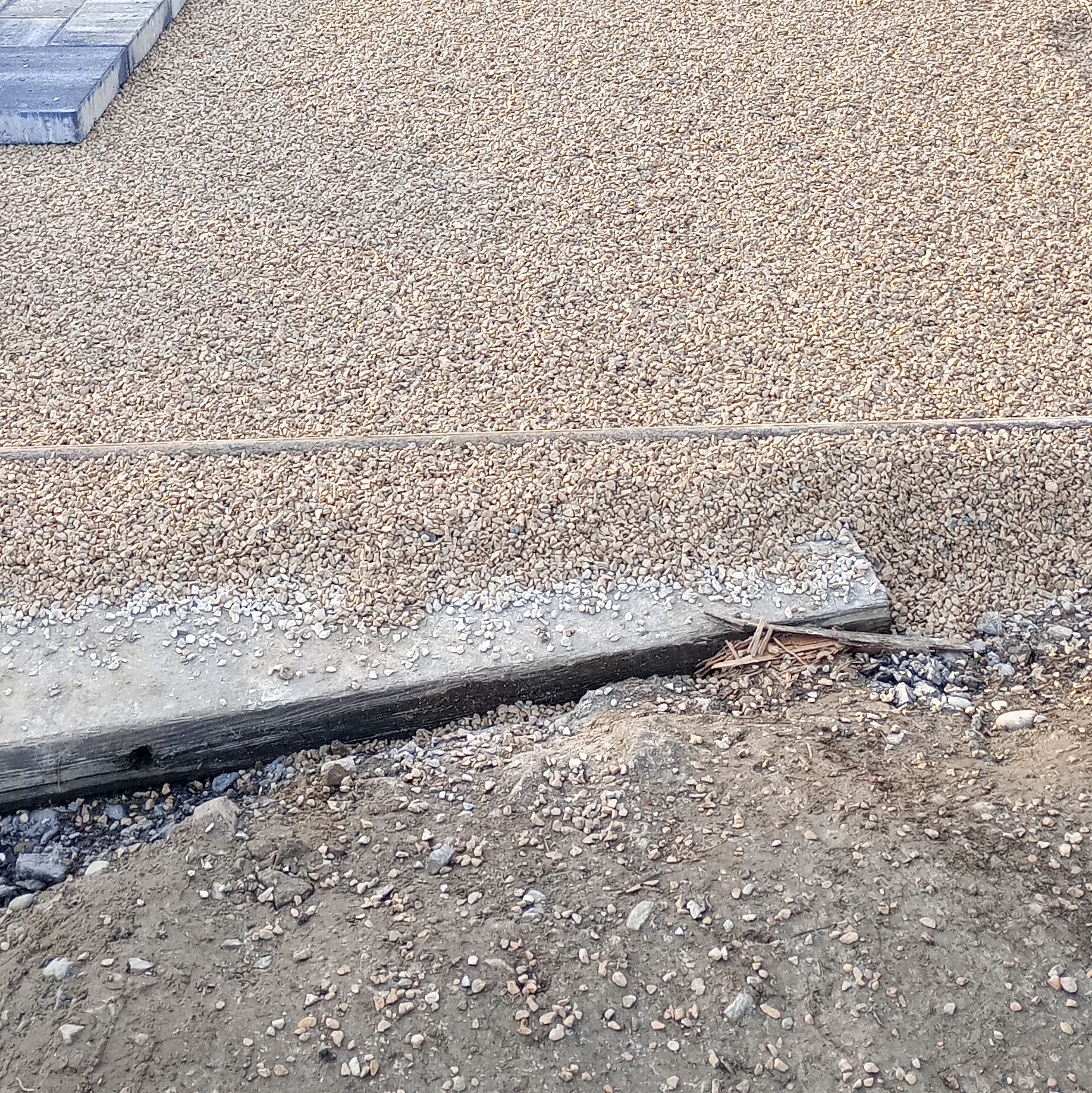 Wyrównany teren z drobnym żwirem, widoczne krawężniki i fragmenty kostki. Przygotowanie podłoża pod kostkę brukową lub inną nawierzchnię. Prace ziemne w toku.