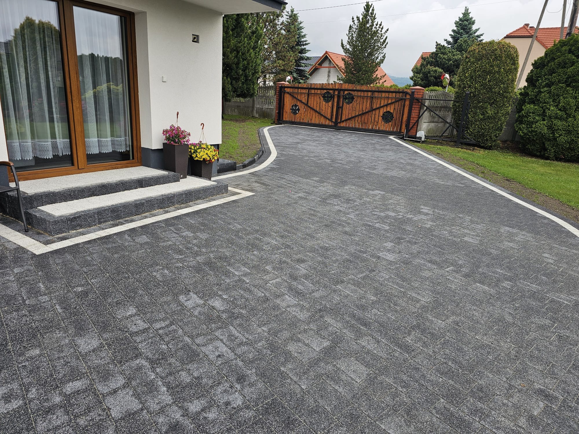 Ułożona, grafitowa kostka brukowa prowadzi od schodów domu do drewnianej bramy wjazdowej. Białe obrzeża podkreślają kształt ścieżki i dodają elegancji.