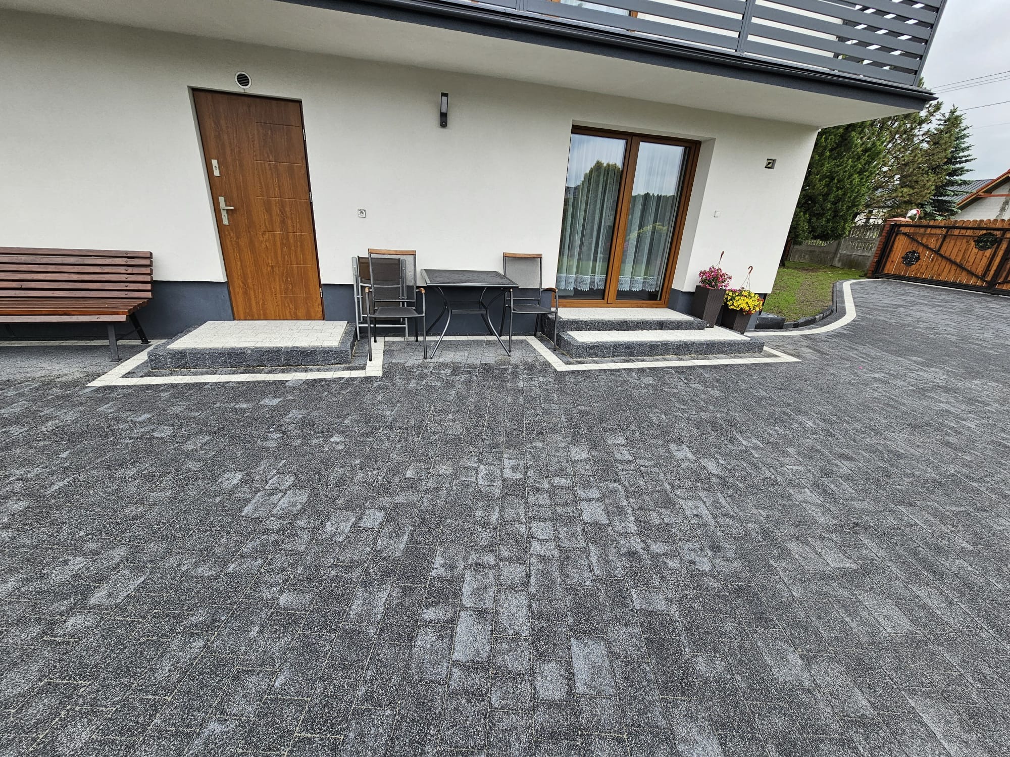 Szara kostka brukowa ułożona przed domem z drewnianymi drzwiami i oknami, uzupełniona o białe obrzeża i schody. Na kostce stoi stolik z krzesłami i ławka.