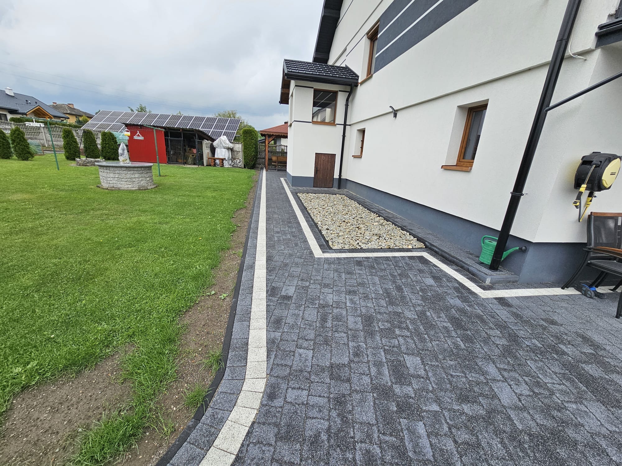 Ułożona, szara kostka brukowa z białymi obrzeżami prowadząca wzdłuż domu. Obok trawnik i studnia. W tle budynek z panelami solarnymi. Pochmurny dzień.