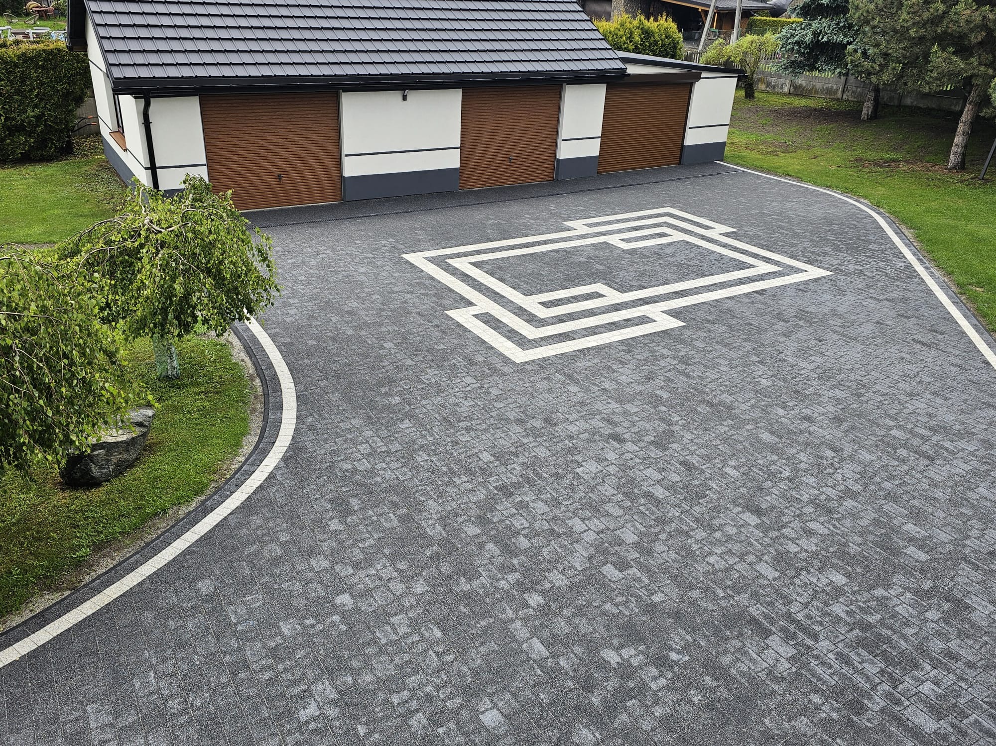 Szara kostka brukowa z białym, geometrycznym wzorem na podjeździe przed budynkiem z trzema garażami. Widok z góry ukazuje estetyczne wykończenie.
