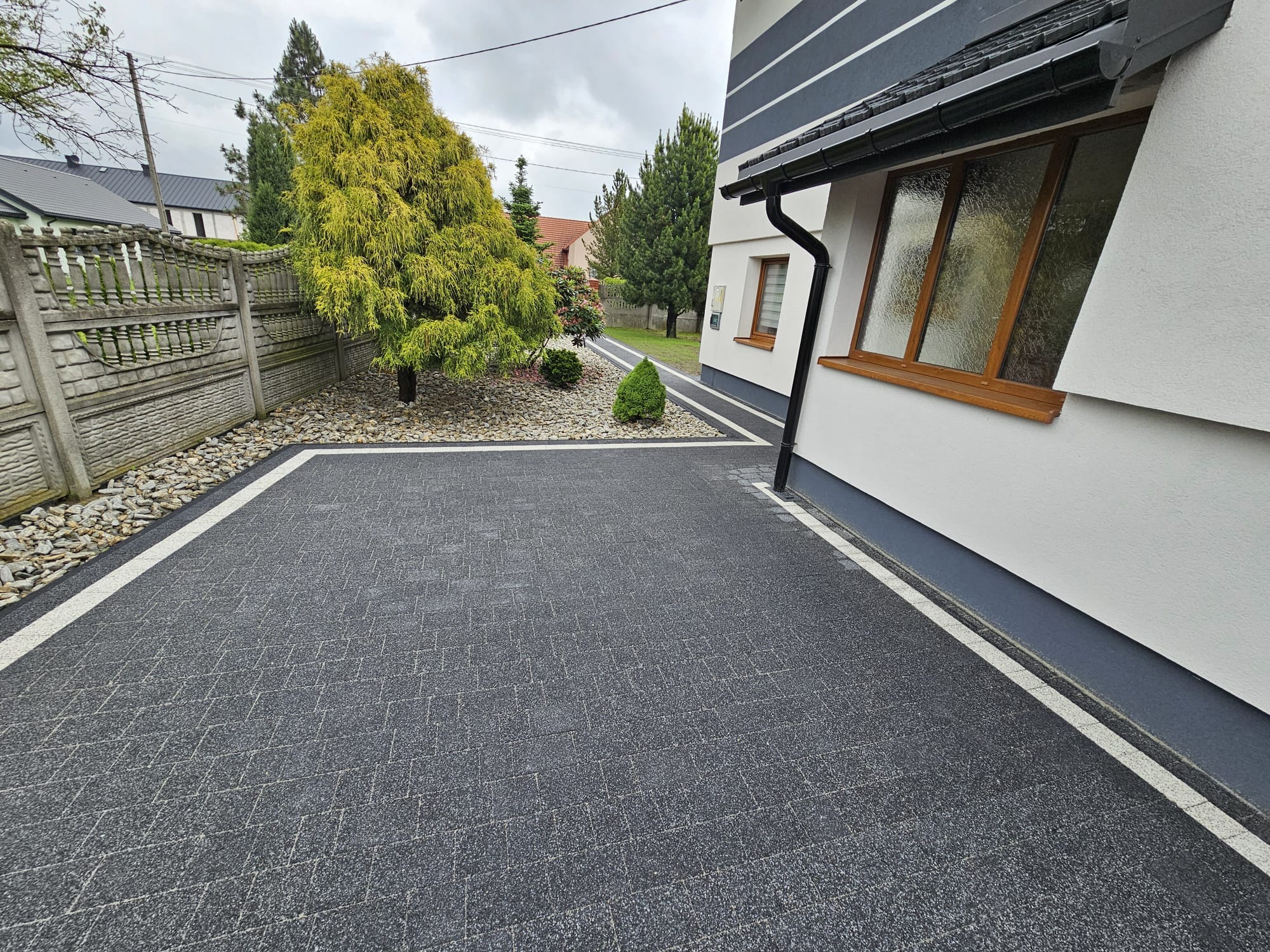 Ciemnoszara kostka brukowa z białymi obrzeżami, ułożona na podjeździe przed nowoczesnym domem z brązowymi oknami. Widoczne kamienie ozdobne i zieleń.