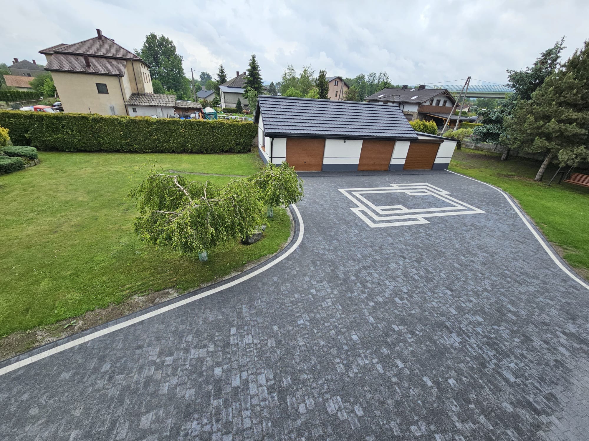 Szara kostka brukowa ułożona na podjeździe z białymi akcentami, widok z góry, w tle budynek garażu z brązowymi bramami i zadbany ogród z zielonym trawnikiem.