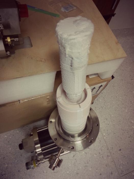 Metalowa kolumna pokryta szronem lub izolacją, prawdopodobnie element urządzenia chłodniczego lub kriogenicznego, z widocznymi przyłączami i elementami konstrukcyjnymi, umieszczona na posadzce.