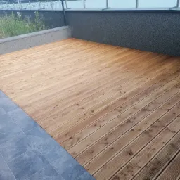 Nowoczesny taras z desek o naturalnym odcieniu drewna, kontrastujący z szarymi płytkami i ciemnoszarym, teksturowanym murem oporowym. Widoczne rośliny za metalową siatką w tle.