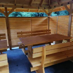 Drewniana altana z ażurowymi ścianami w Jedlni Letnisko, wyposażona w stół i ławy z drewna, widok na zieleń z zewnątrz.