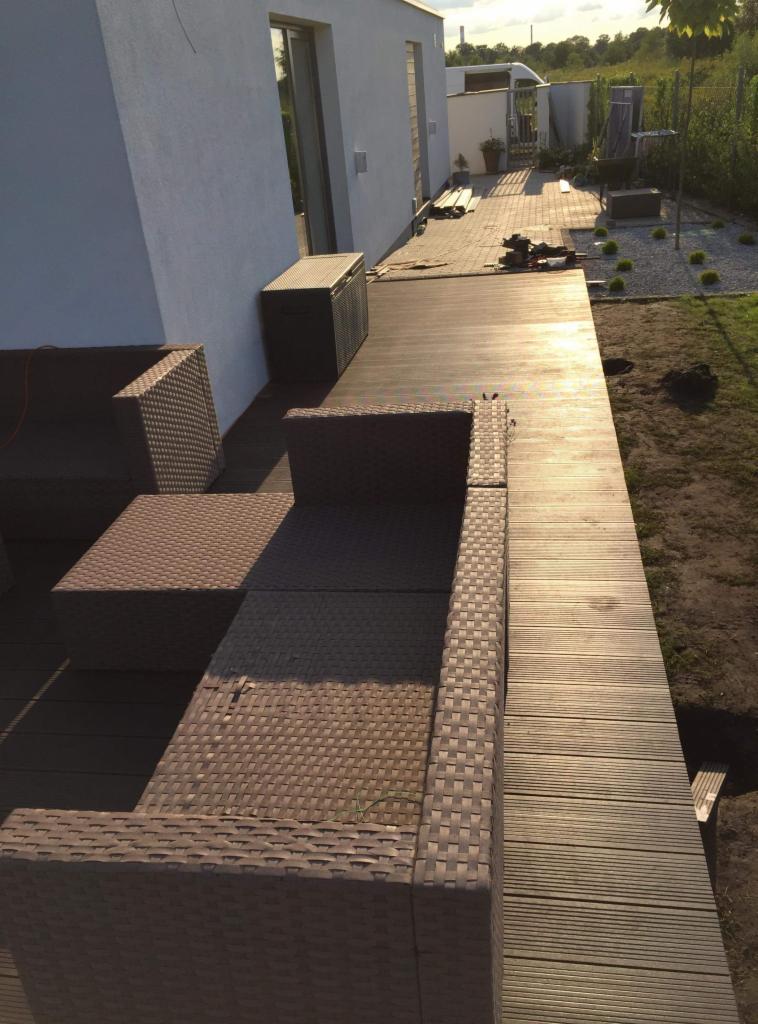 Wykończony taras z desek w odcieniach brązu, z kompletem mebli ogrodowych z rattanu, skrzynią do przechowywania i widokiem na ogród z kamieniami i roślinami.