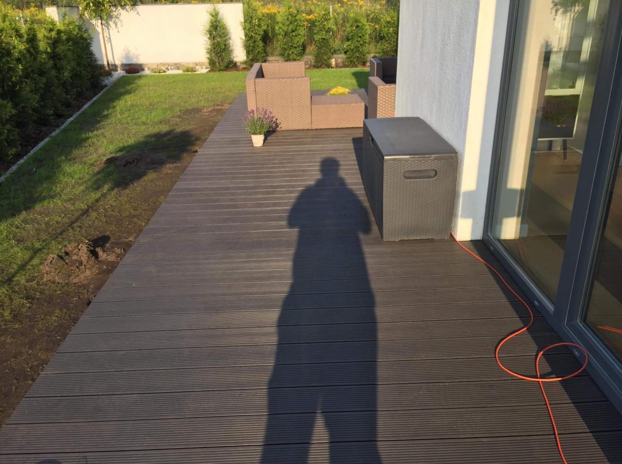 Ciemnoszary taras z desek kompozytowych, na którym stoi rattanowy komplet wypoczynkowy i skrzynia, cień fotografa na deskach, obok domu z dużym oknem.