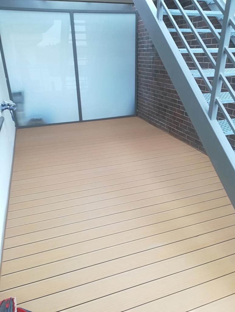 Jasny taras kompozytowy z subtelnym rysunkiem drewna, wykonany w Jedlni Letnisko, przylegający do schodów zewnętrznych i ściany z cegły klinkierowej.