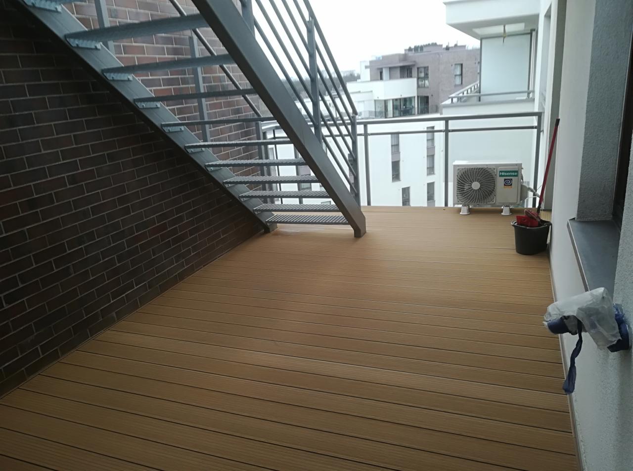 Zewnętrzny taras z desek kompozytowych, z metalowymi schodami prowadzącymi na górę. Na tarasie widoczny klimatyzator, wiadro i mop przy ścianie. W tle budynki mieszkalne.