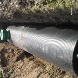 Czarny zbiornik z zieloną pokrywą, częściowo zakopany w ziemi w wykopie, widoczne fragmenty trawy i ziemi. Zbiornik z wyraźną fakturą, ułożony poziomo.