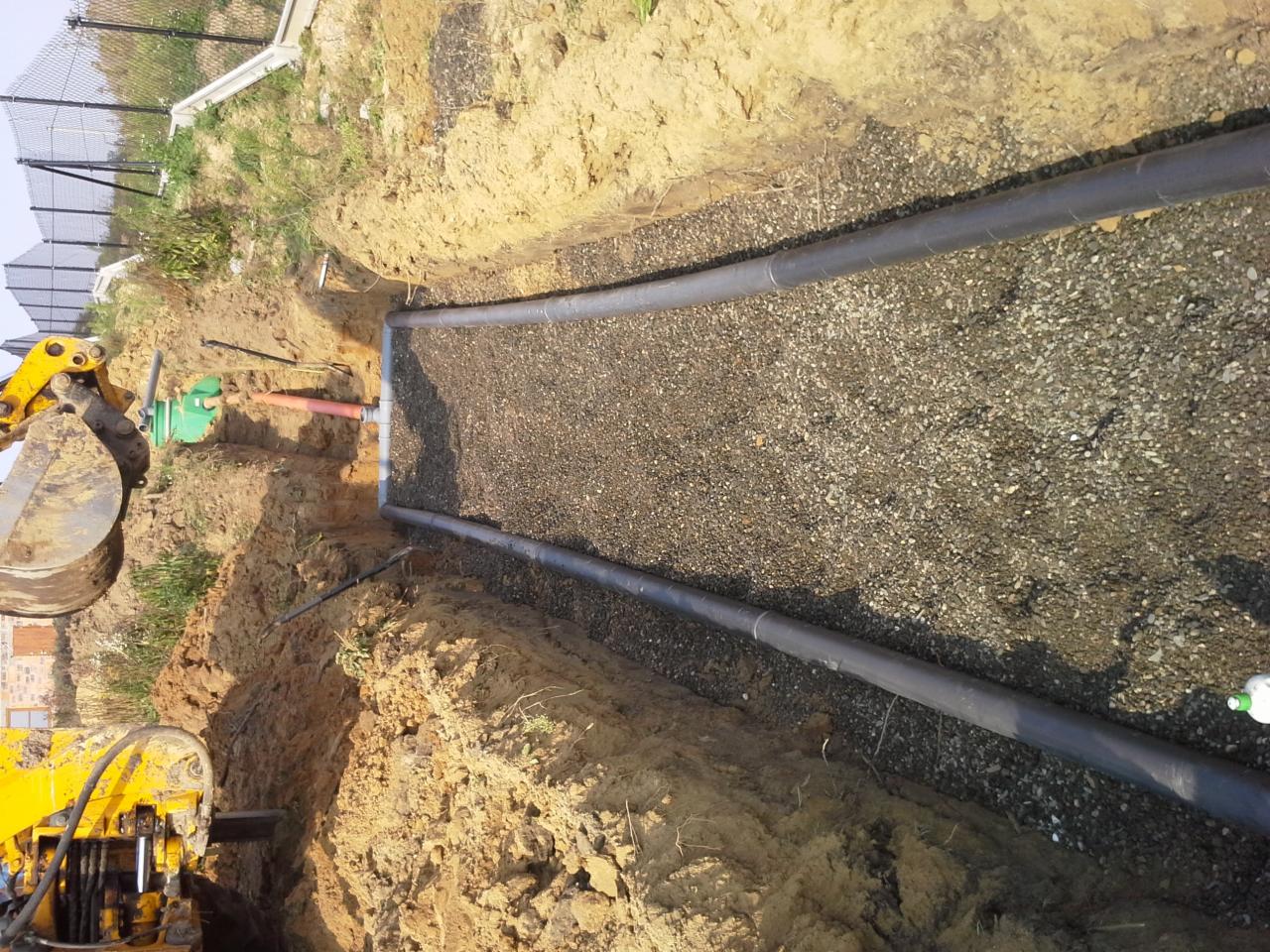 Wykop z ułożonymi rurami i żwirem, prawdopodobnie drenaż. Żółta koparka w tle. Widoczne połączenia rur. Ujęcie z góry, lekko obrócone.