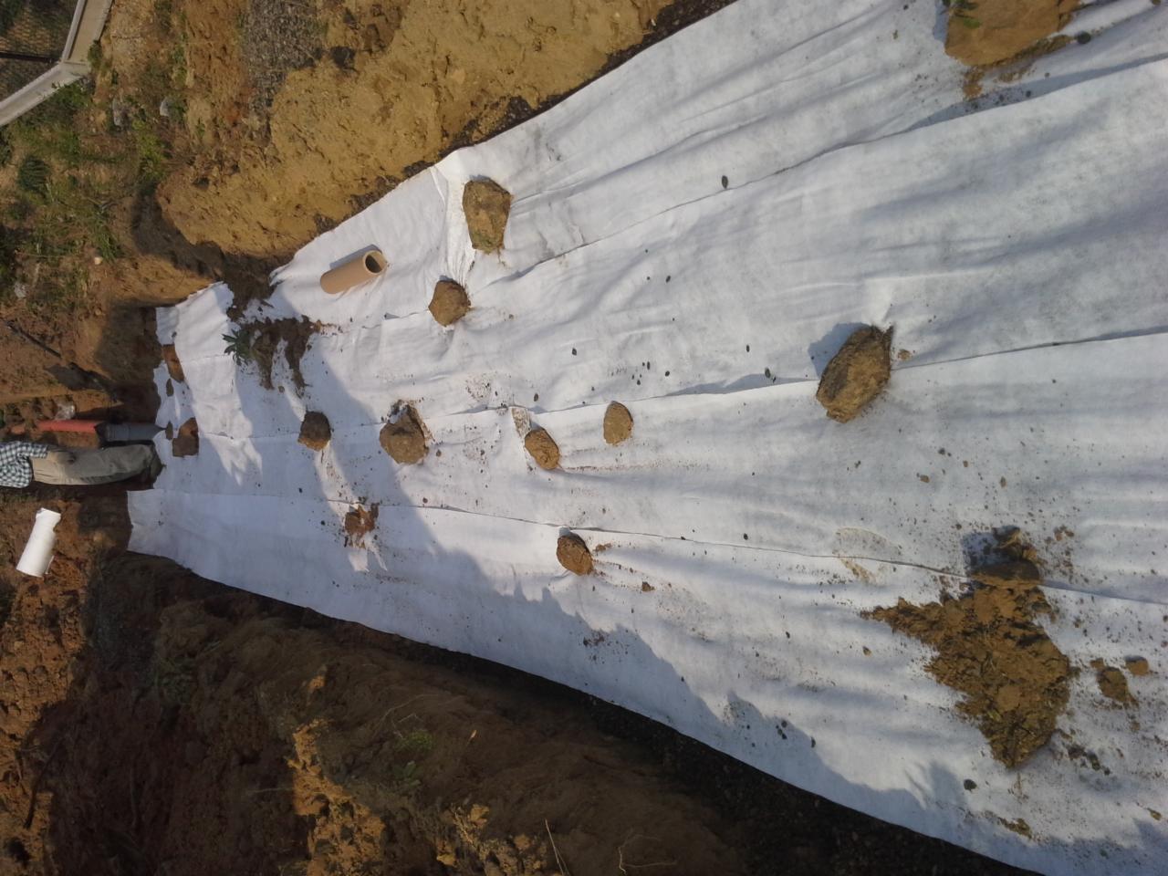 Wykop z rurami drenażowymi, przykryty białą geowłókniną i obciążony ziemią. Elementy instalacji przydomowej oczyszczalni ścieków w trakcie budowy.