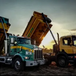 Dwa wywrotki: zielono-żółty Kenworth i żółta Scania, z podniesionymi skrzyniami ładunkowymi, na placu budowy podczas zachodu słońca.