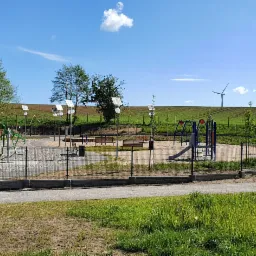 Nowoczesny plac zabaw z urządzeniami rekreacyjnymi i oświetleniem solarnym, otoczony ogrodzeniem, na tle zielonych pól i turbin wiatrowych.