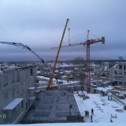 Zimowy widok z góry na plac budowy z dwoma żurawiami, pompą do betonu i pracownikami w odblaskowych kamizelkach układającymi zbrojenie pod wylewkę fundamentów.