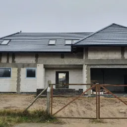 Niedokończony dom jednorodzinny z szarej dachówki, widoczne bloczki betonowe, okna z szarymi ramami, brama wjazdowa i żółty znak drogowy.