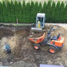 Wykopany dół pod basen w ogrodzie, z widocznym pracownikiem w dole, mini koparką i wywrotką na powierzchni.