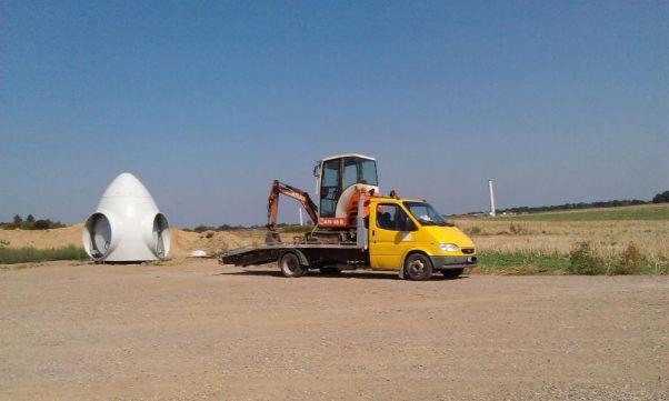 Żółty samochód dostawczy z pomarańczową mini koparką na platformie transportuje element turbiny wiatrowej na placu budowy z widocznym w tle masztem turbiny.