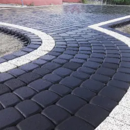 Ułożona nawierzchnia z czarnej kostki brukowej z białym obramowaniem, widoczne elementy drenażu liniowego i podsypka piaskowa.