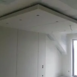 Wnętrze w trakcie prac wykończeniowych: widoczna konstrukcja sufitu podwieszanego z wbudowanym oświetleniem punktowym oraz ściany przygotowane do malowania, widoczne łączenia płyt na suficie...