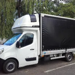 Biały samochód dostawczy Renault Master z czarną plandeką, zaparkowany pod drzewem, widoczny z boku, z nadbudową kabiny sypialnej.