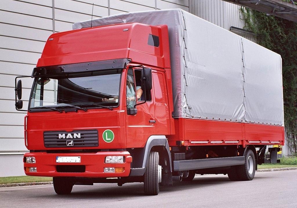 Czerwona ciężarówka MAN z plandeką na tle budynku o jasnej elewacji i zieleni. Widoczna kierowca w kabinie.