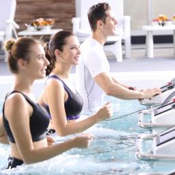 VITAMELA Sp. z o.o. - Trzy osoby ćwiczą na bieżniach zanurzonych w basenie, widoczne z profilu, w tle stolik z owocami i białe krzesła.
