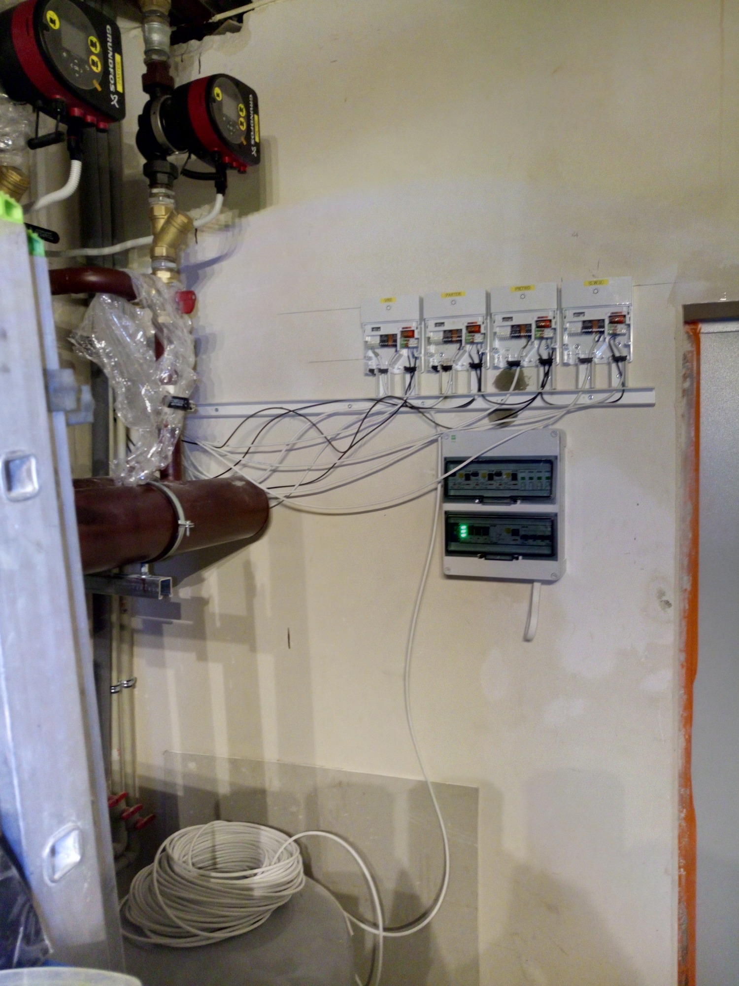 Węzeł cieplny w piwnicy z dwoma pompami obiegowymi Grundfos, rurami, przewodami elektrycznymi i rozdzielnicą na ścianie, obok stoi drabina.