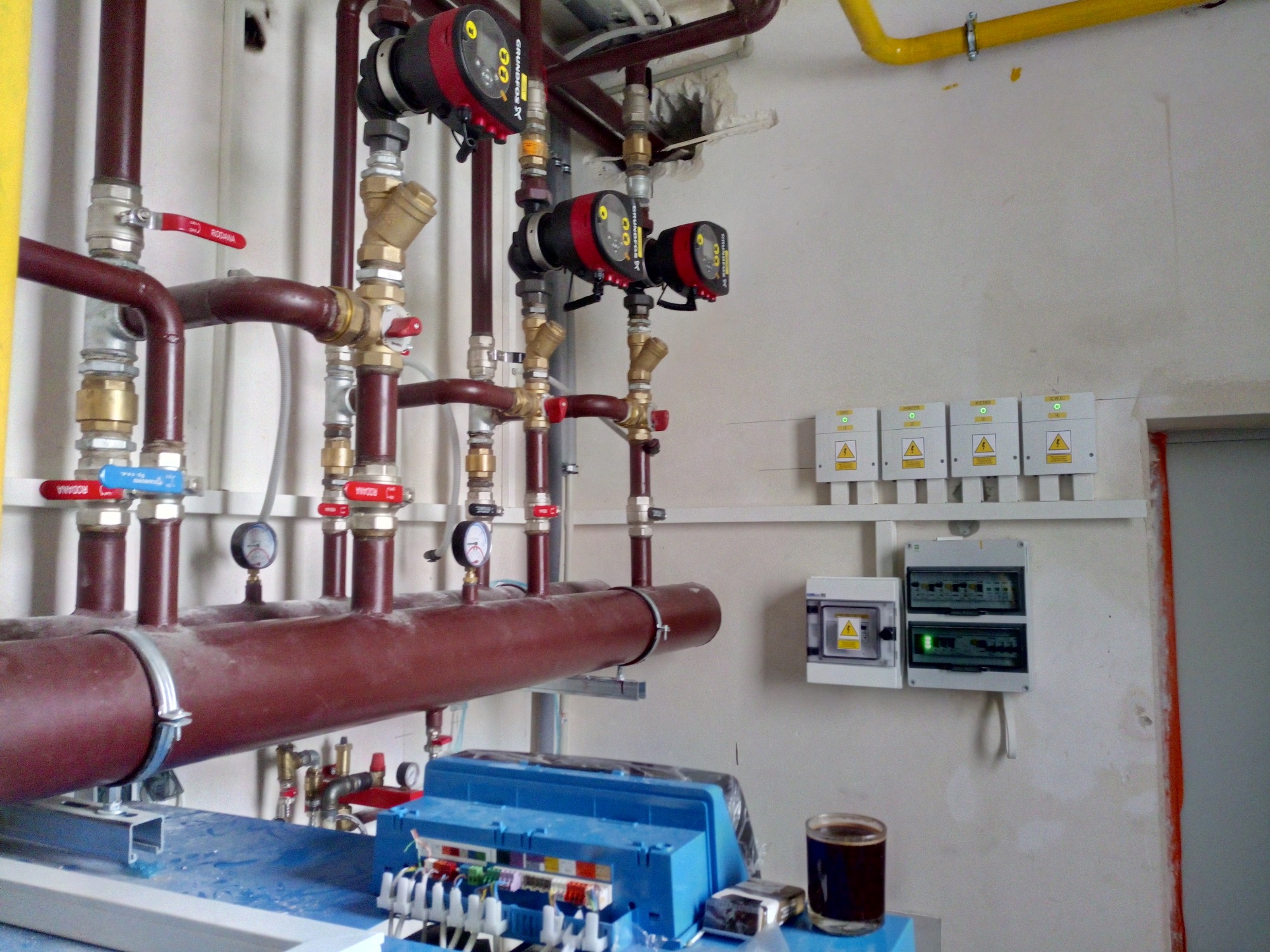 Instalacja grzewcza z brązowymi rurami, zaworami z czerwonymi uchwytami, trzema pompami obiegowymi Grundfos, manometrami, żółtą rurą gazową pod sufitem i skrzynkami elektrycznymi na ścianie.