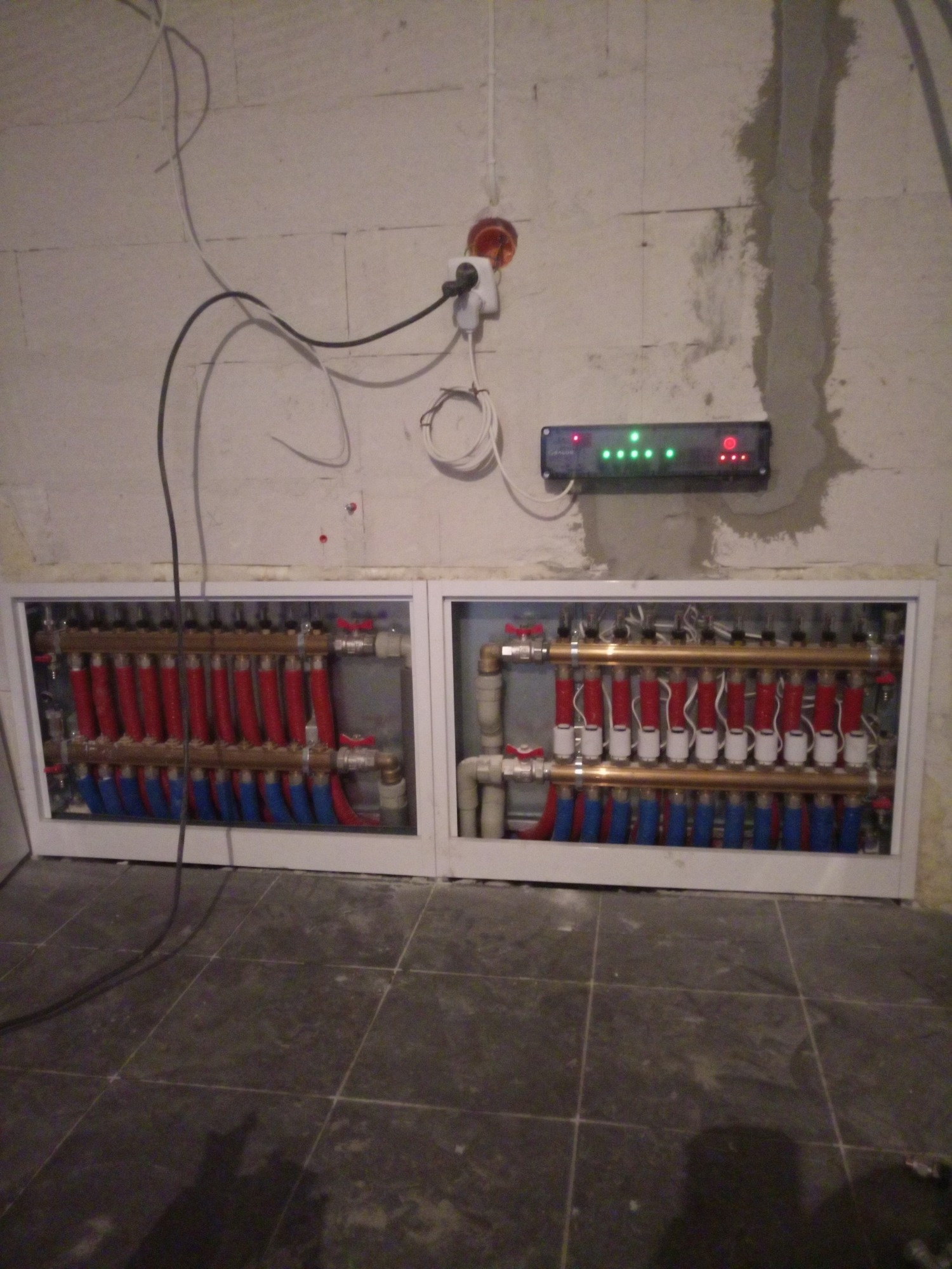 Rozdzielacz ogrzewania podłogowego z czerwonymi i niebieskimi rurami w białej zabudowie, z podłączonym sterownikiem elektronicznym z diodami LED na surowej ścianie z betonu komórkowego.