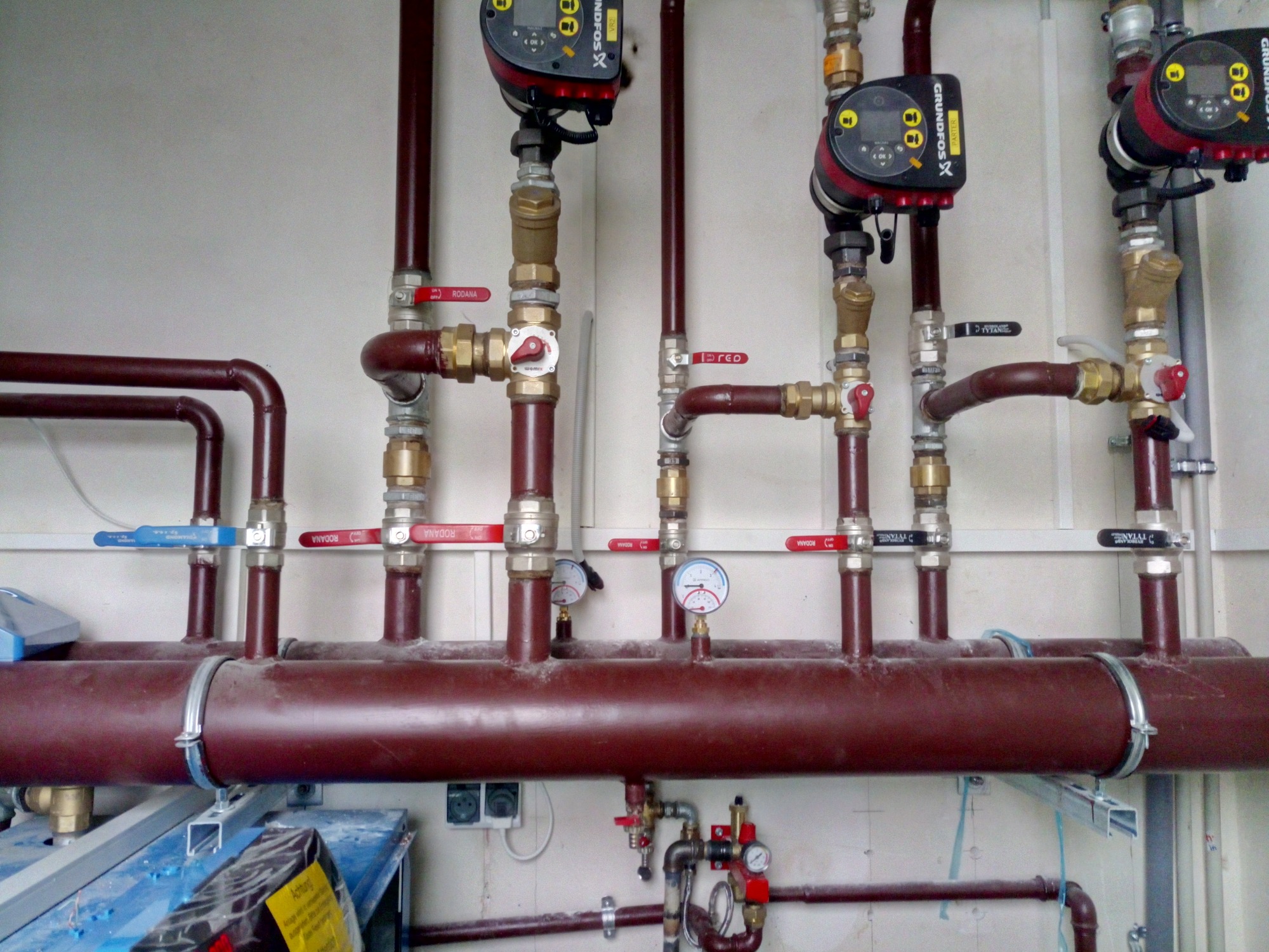 Instalacja centralnego ogrzewania z brązowymi rurami, zaworami oznaczonymi kolorami i pompami obiegowymi Grundfos zamontowanymi na ścianie w pomieszczeniu technicznym.